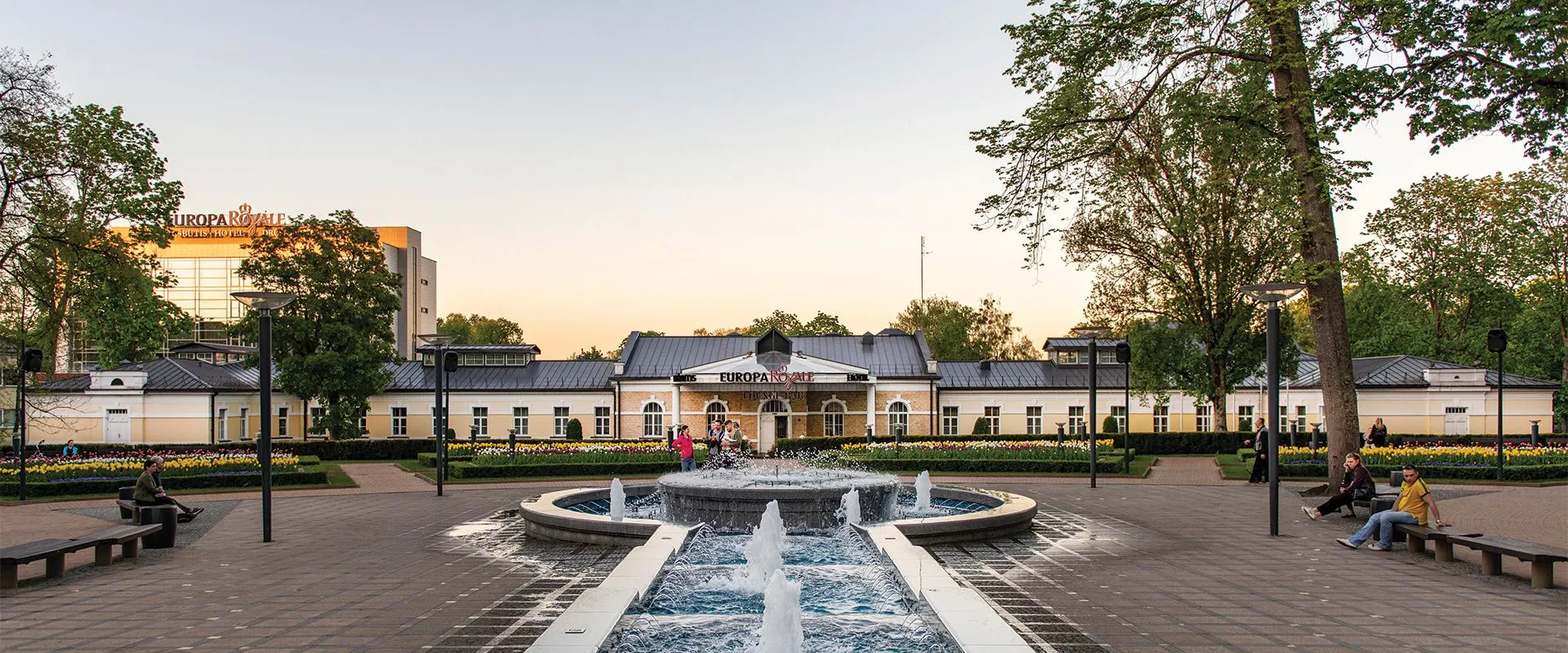 Fountain and tree-lined promenade in front of Europa Royale Druskininkai at dusk