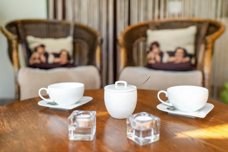 White teacups, a sugar bowl and glass tealight holders on a wooden table beside wicker armchairs