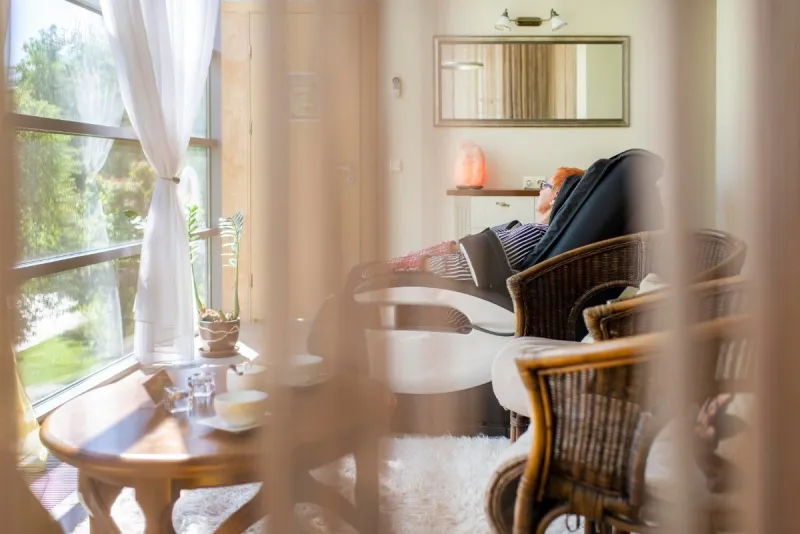 View past a sheer curtain into a sitting corner with wicker armchairs, a round table and a wall mirror