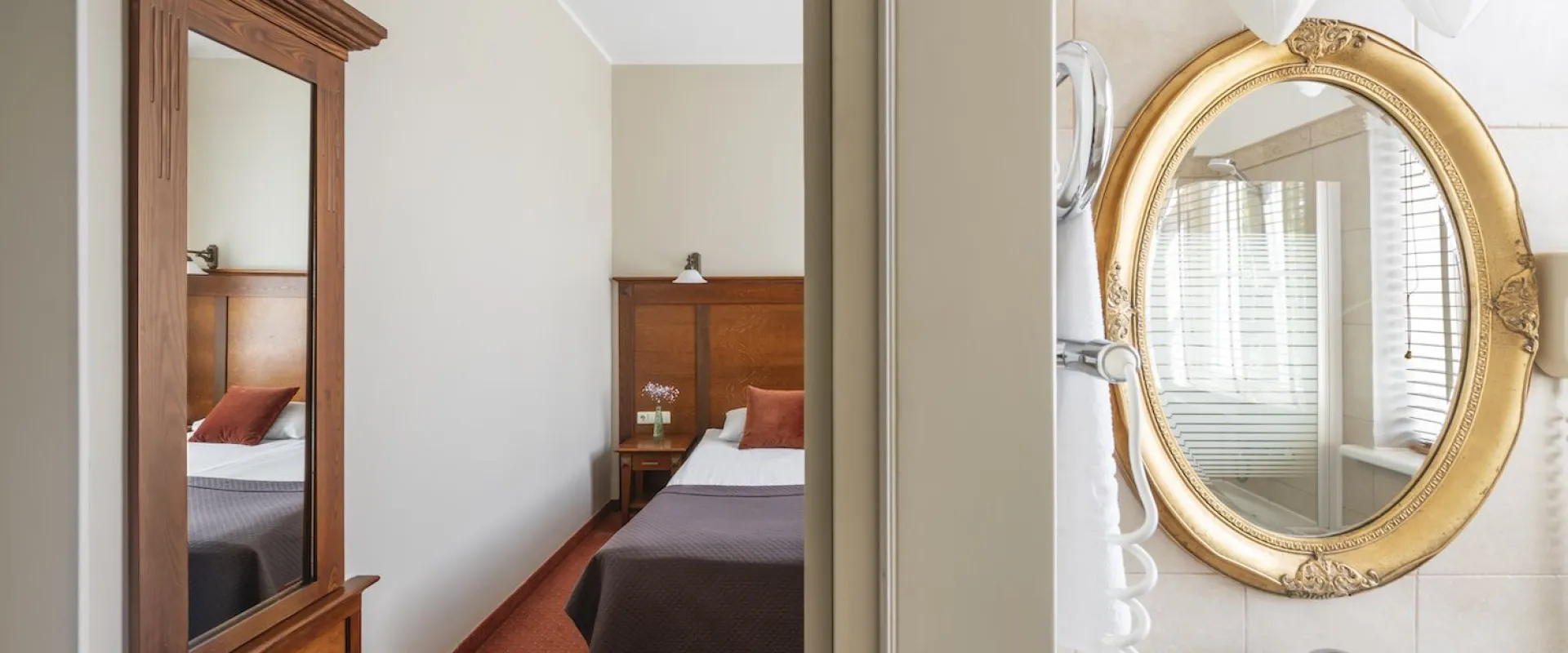 View past a tall mirror reflecting a bed, into a bathroom with a gilded oval mirror above the basin