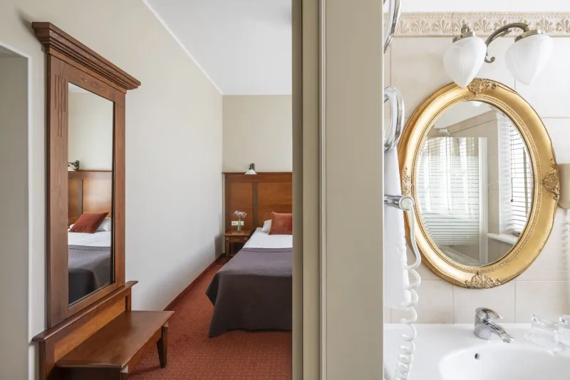 View past a tall mirror reflecting a bed, into a bathroom with a gilded oval mirror above the basin