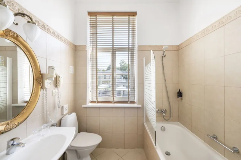 Beige-tiled bathroom with a bath and shower, toilet, basin and gilded mirror beside a shuttered window