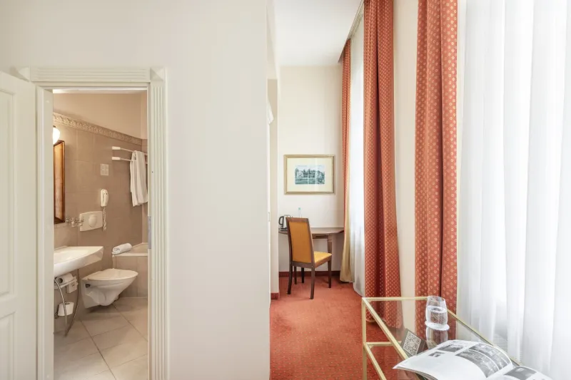 View from a room's entrance past the open en-suite bathroom to a desk and red curtained window