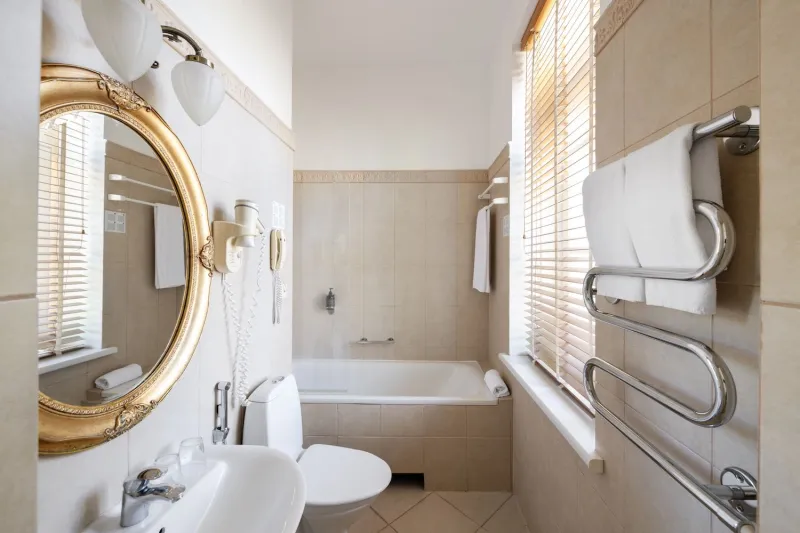 Bathroom with a bathtub, oval gilded mirror above the basin and a chrome heated towel rail by a blinded window