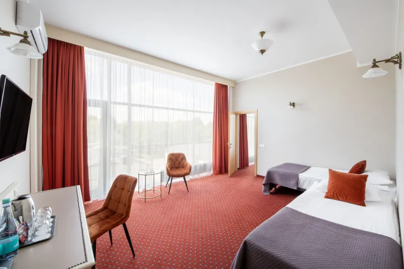 Room with two chairs and a round table at a wide window overlooking trees, twin beds alongside