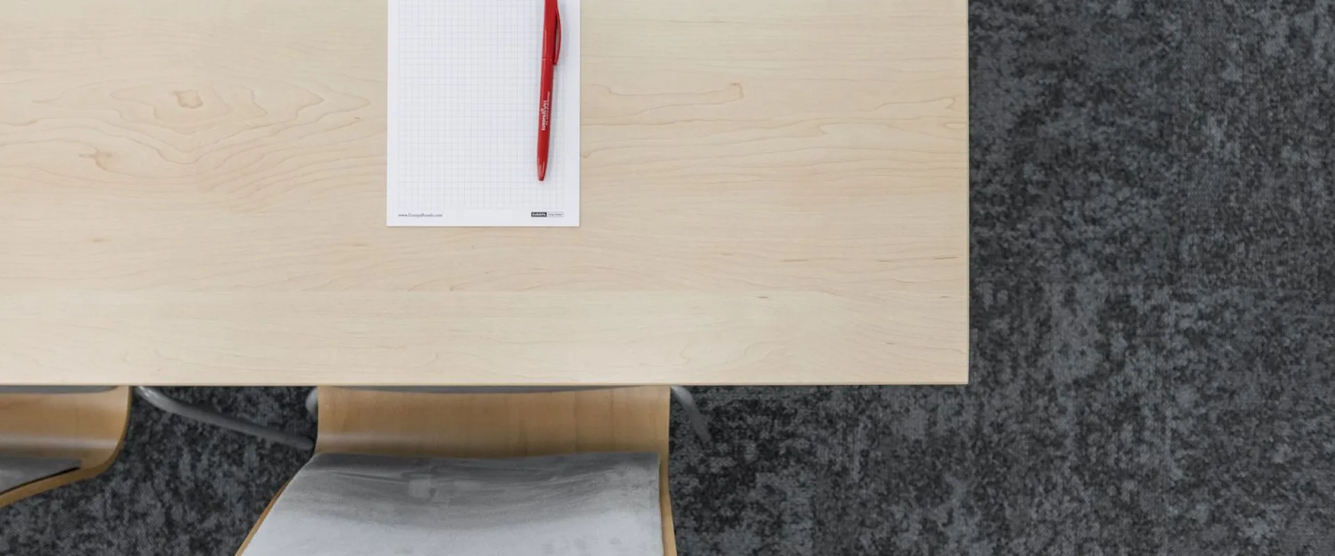 Notepad and red pen on a light wooden conference table above a grey stone floor