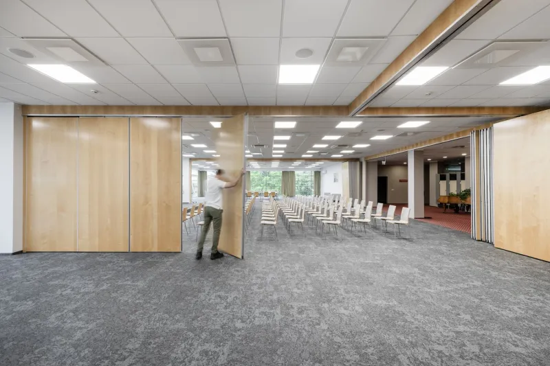 Person sliding a wooden partition to divide a conference room set with rows of chairs
