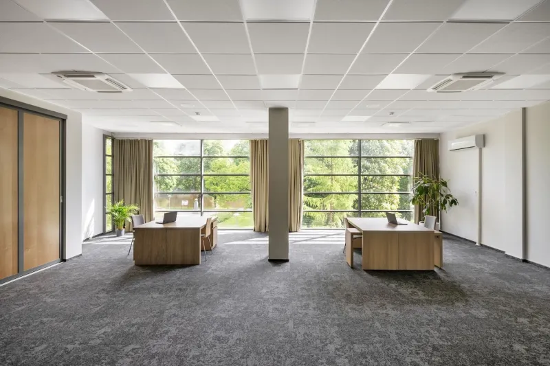 Empty conference room with two wooden desks and laptops facing full-height windows onto trees