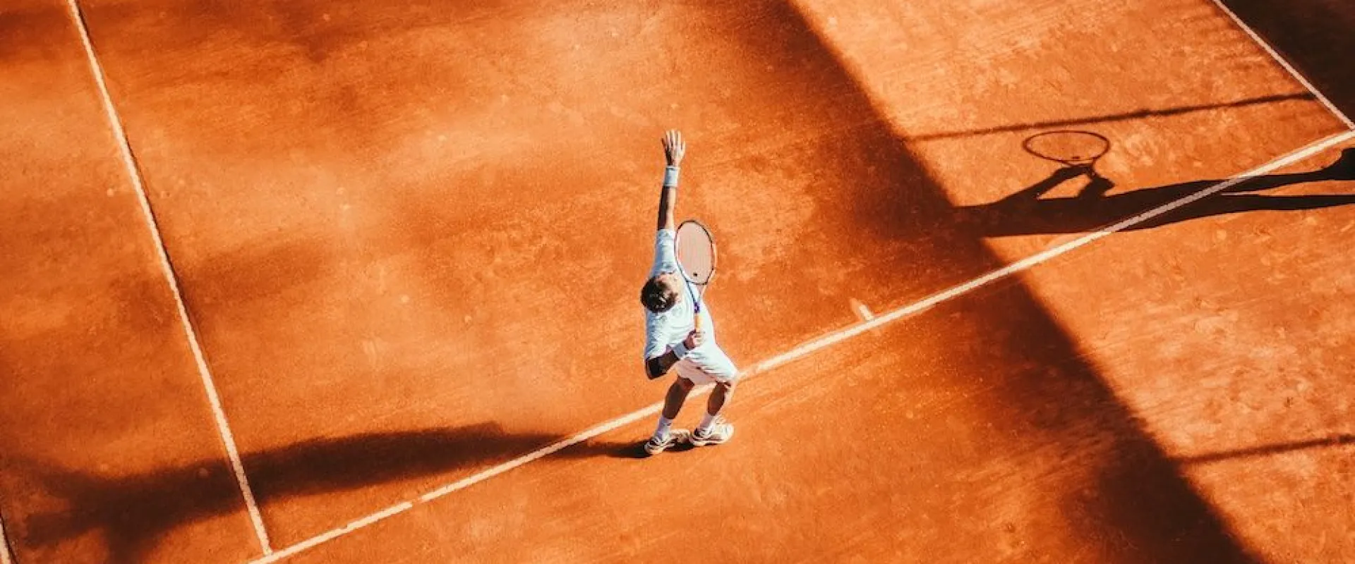 Tennis player serving on a clay court in low sunlight