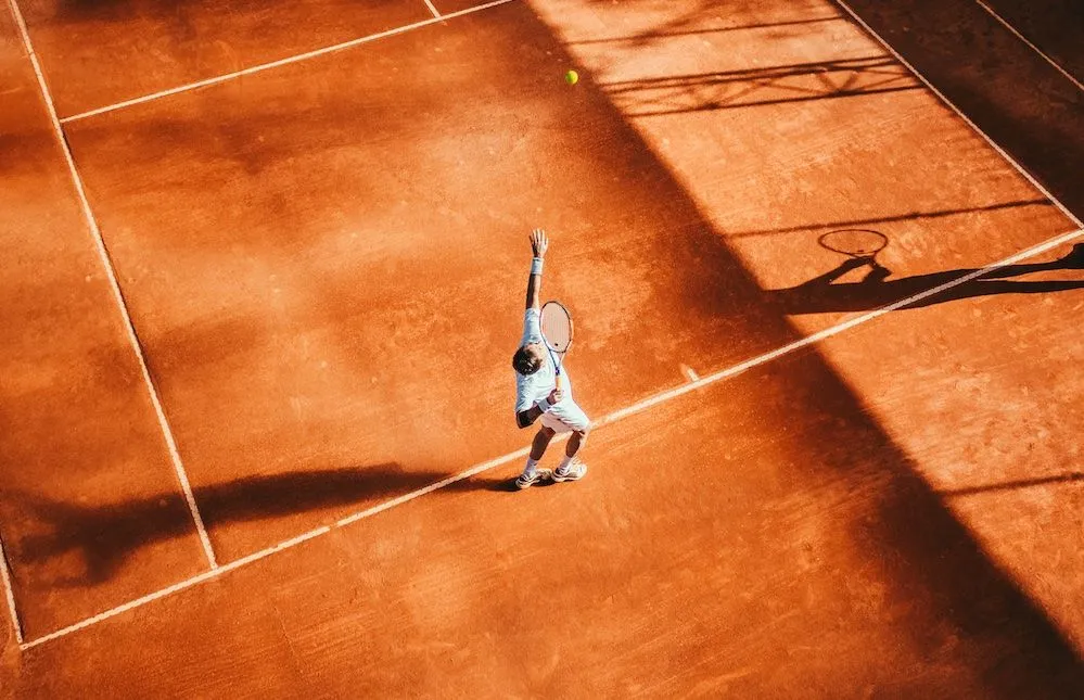 Tennis player serving on a clay court in low sunlight