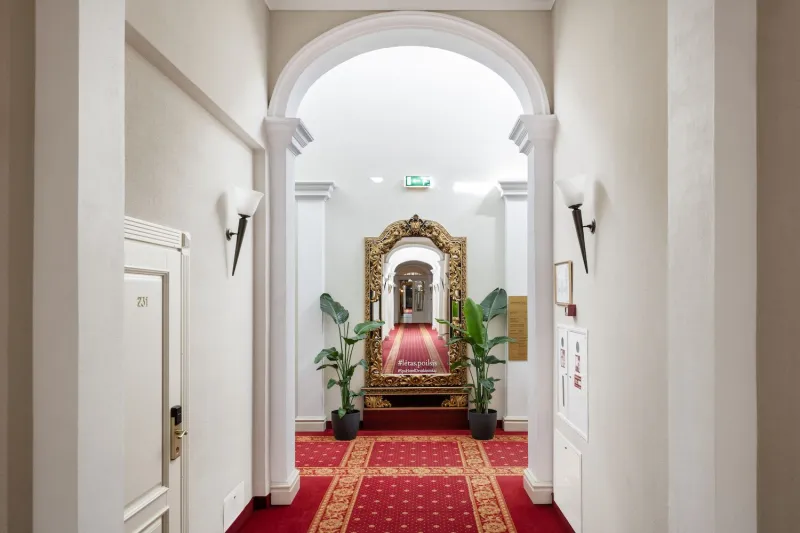 Arched hotel corridor with a red patterned carpet leading to a gilded mirror