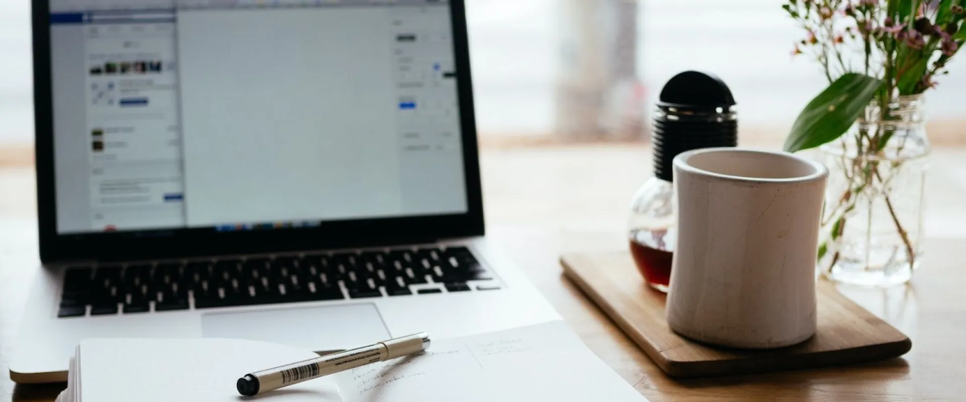 Laptop, open notebook and a cup of coffee on a desk by the window